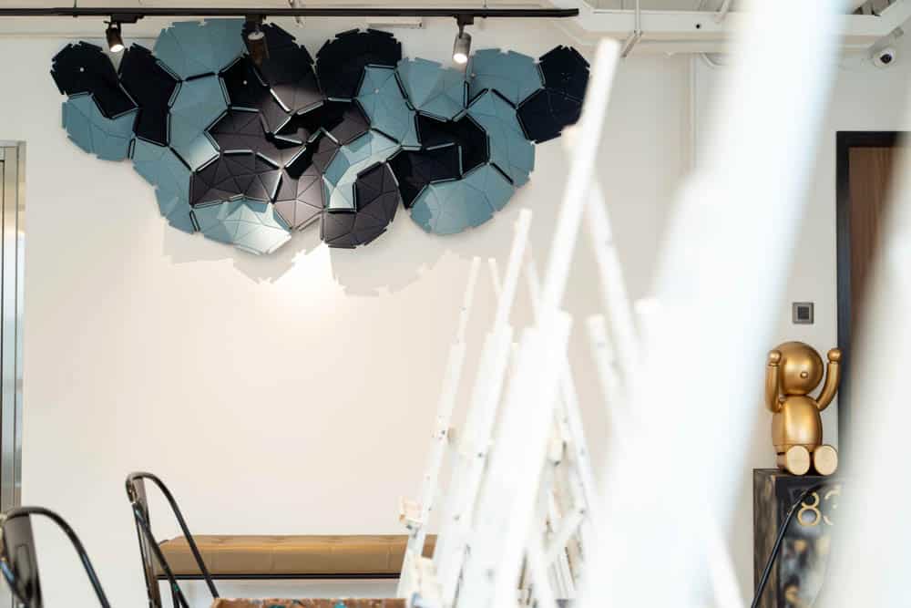 A modern interior features a wall art installation of blue and black geometric shapes resembling umbrellas, reminiscent of an abstract oil painting. In the foreground, chairs have diagonal white cables, and a small golden sculpture adorns a side table.