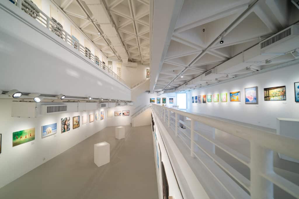 A spacious, modern art gallery with white walls and ceilings, displaying colorful paintings on both upper and lower levels, with railings and bright lighting throughout.
