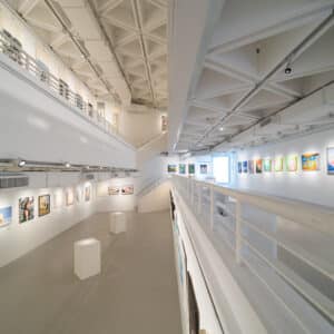 A spacious, modern art gallery with white walls and ceilings, displaying colorful paintings on both upper and lower levels, with railings and bright lighting throughout.
