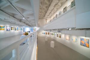 A spacious, modern art gallery with white walls and high ceilings displays colorful paintings from a recent art competition along both levels. Two white benches sit on the floor below, brightly lit by overhead lights.