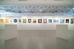 A modern art gallery with a white interior displays colorful paintings from an art competition in a single row along the back wall. Two empty white display stands are positioned in the center of the spacious, well-lit room.