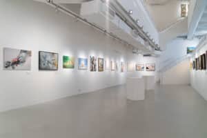 A modern art gallery with white walls displays various colorful paintings from an art competition. The spacious room has a light gray floor, spotlights above, and a staircase at the back. Two white display pedestals stand in the center.