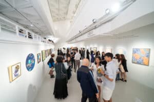 A bright, modern art gallery filled with people viewing and discussing colorful paintings on white walls during an art competition. The diverse, engaged crowd adds excitement to the open, lively space with high ceilings and track lighting.